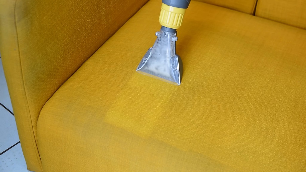 Mlaznica dubinskog usisivača prska i čisti žutu tkaninu na sofi.